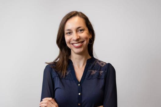 Lizzie is smiling at the camera. She is wearing a blue top, folding her arms and has brown hair. 