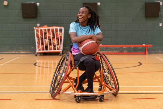A black young woman in a wheelchair is laughing and holding a basketball