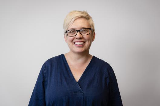 Heather smiling at the camera. She's a white woman with short blonde hair, wearing a black rimmed glasses