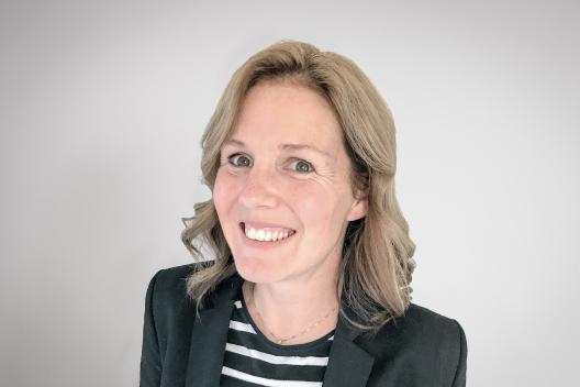 Zoe smiling at the camera. She's a white woman with shoulder length blonde hair, wearing a black blazer and stripy top