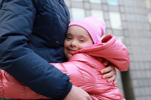 A girl in a pink hat and coat hugs an adult in blue.