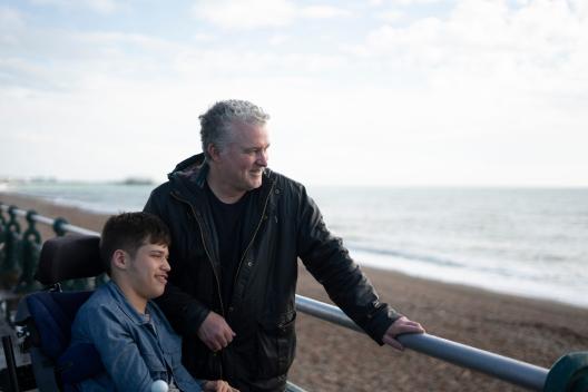 A young man in a wheel chair and an older man standing looking out at the sea