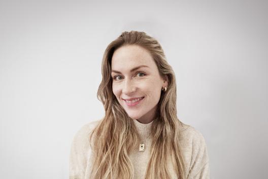 Alexandra smiling at the camera. She's a white woman with long blonde hair, wearing a beige turtle neck jumper and gold earrings.