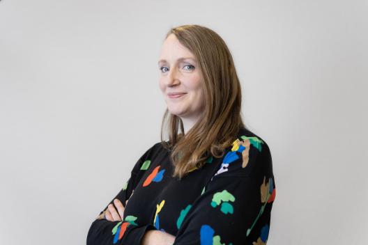 Anna smiling at the camera. She's a white woman with blue eyes and dark blonde hair, wearing a black jumper with colourful shapes on it