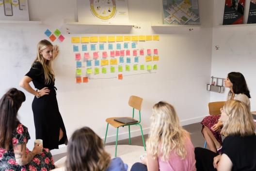 A woman standing by a wall of post it notes with a small audience