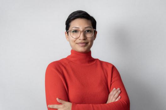 Jahaan smiling at the camera. She's a woman of colour with short brown hair, wearing a red turtleneck jumper and glasses