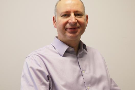 James smiling at the camera. He's a white man with short grey hair, wearing a lilac shirt.