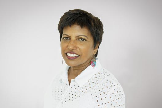Radhika smiling at the camera. She's a woman of colour with short brown hair, wearing a white shirt and pink and blue earrings