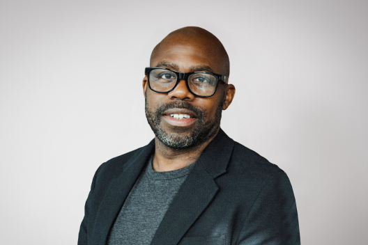 Shane smiling at the camera. He's a bald black man with a short beard, wearing black rimmed glasses with a grey t-shirt and a black jacket