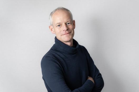 Tom stands with his arms crossed looking at the camera. He's a middle aged white man with blue eyes and short grey hair. He wears a blue jumper