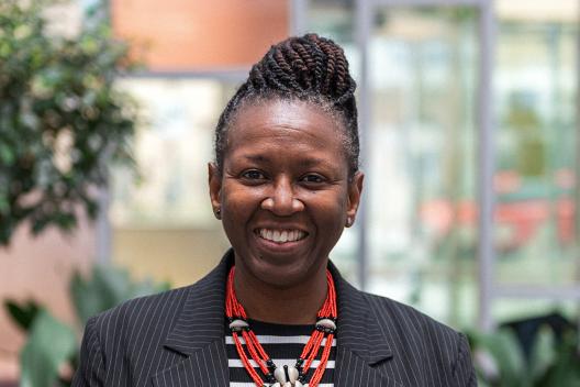 Sola smiling at the camera. She's a black woman with braided hair tied up. She is wearing a red necklace and a pinstriped jacket.
