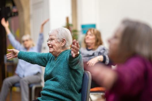 A smiling older woman sat on a chair holding her arms up in the air in an exercise class