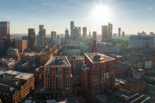 Aerial view of Manchester, UK, on a sunny day, featuring skyscrapers, construction cranes, historic brick buildings, and modern architecture.