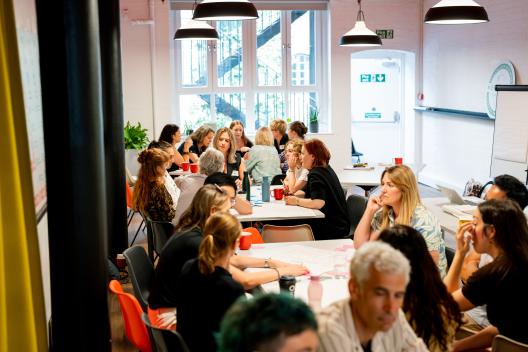 A room full of people separated into groups, each group is gathered around a table where they are in conversation.  