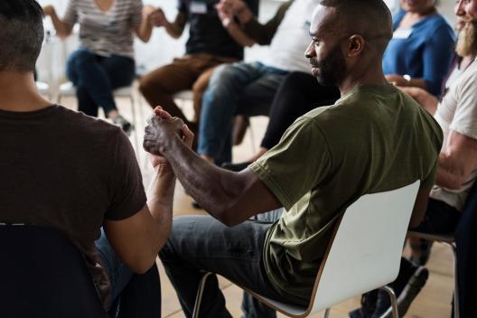 A group of men and women are sat on chairs in a circle holding hands, they are wearing name tags and are looking at each other. 