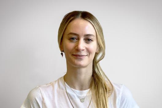 A woman wearing a white t-shirt and blonde hair is smiling into the camera. Behind her is a white wall.  