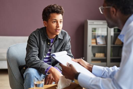 A teenage boy wearing a striped shirt, jacket and jeans is looking across a table at an older man in a shirt. The man is holding a clipboard and a pen, he is engaging in deep conversation with the teenage boy. On the table between them is a box of tissues and a glass of water.  