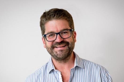 Matthew Horne smiles at the camera, he is wearing a blue striped shirt and glasses