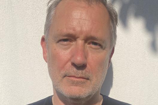 A man wearing a black t-shirt is looking directly at the camera, he has a grey beard and grey hair
