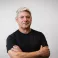Mike stands with his arms crossed looking at the camera. He's a middle aged white man with blue eyes and short grey hair. He wears a black t-shirt.