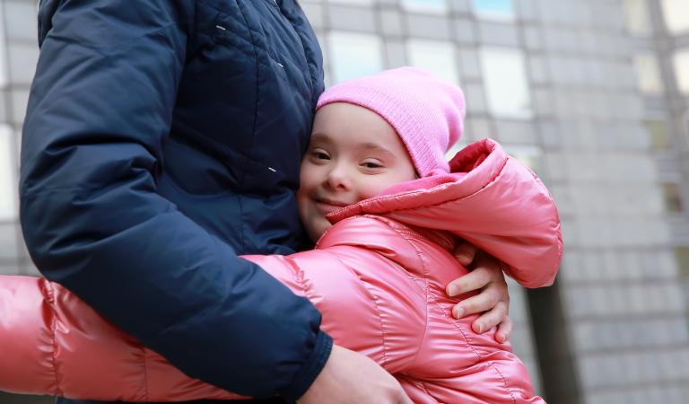 A girl in a pink hat and coat hugs an adult in blue.