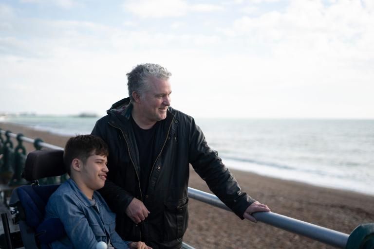 A young man in a wheel chair and an older man standing looking out at the sea