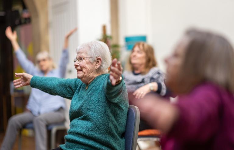 A smiling older woman sat on a chair holding her arms up in the air in an exercise class