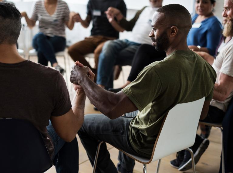 A group of men and women are sat on chairs in a circle holding hands, they are wearing name tags and are looking at each other.