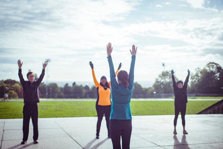 A fitness instructor leads a small group of people in an outdoor exercise class, all with their arms raised overhead.