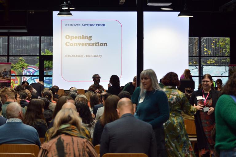 A large indoor crowd sits in rows of wooden chairs, facing a massive projection screen at the front of a room with an industrial aesthetic. The screen displays the text "CLIMATE ACTION FUND" followed by "Opening Conversation" and "9:45-10.45am - The Canopy" inside a rounded pink border. Several people are standing and talking near the front and in the aisles. Large windows in the background reveal an outdoor area with graffiti art and green foliage.