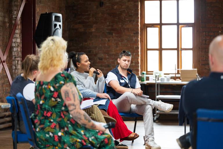 A panel of people sitting in a circle during a meeting or event in a room with exposed brick walls and large windows. In the center, a woman with dark hair up in a bun speaks into a microphone while wearing a grey sweater. Beside her, a man with sunglasses on his head listens with a notebook on his lap. In the foreground, other participants listen attentively.