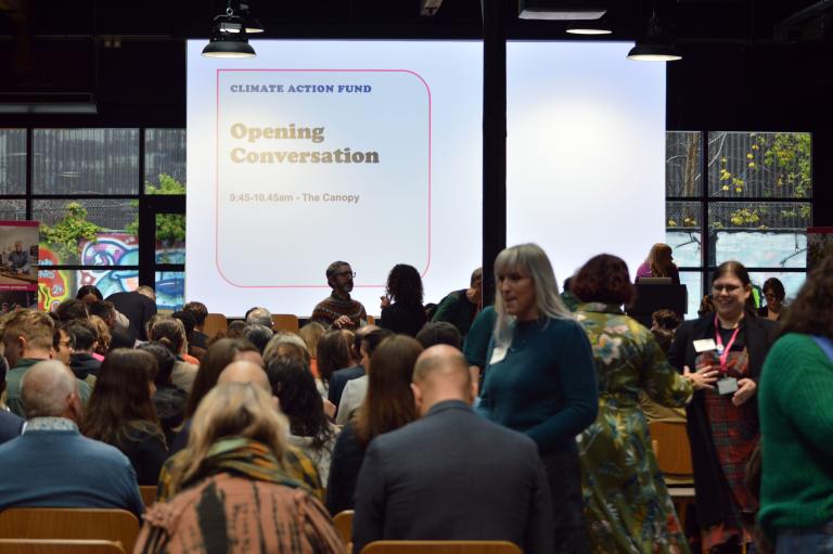 A crowded indoor conference room filled with seated attendees viewed from behind, facing a large presentation screen. The screen reads "Climate Action Fund: Opening Conversation, 9:45–10:45am – The Canopy." People are chatting and moving around in the foreground and aisles before the presentation starts.