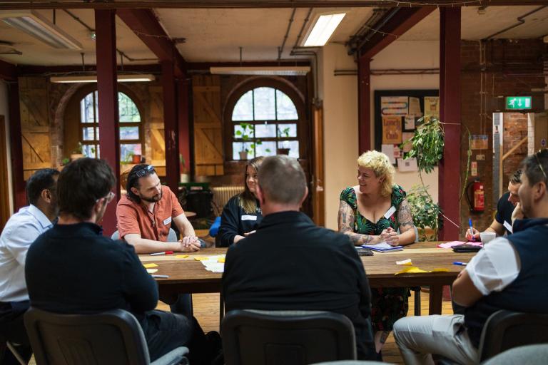 A group of people gathered around a large wooden table during a collaborative workshop, with sticky notes and pens spread across the workspace.