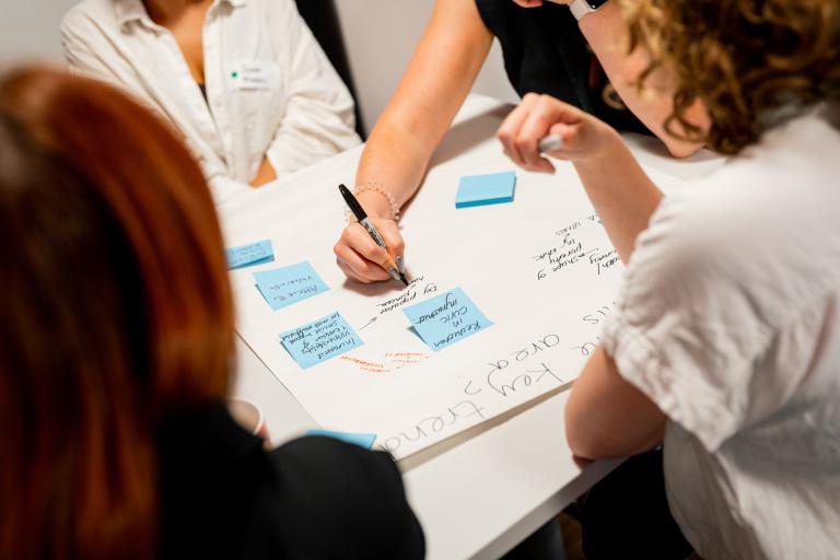 Close-up view of workshop participants gathered around a table, placing blue sticky notes and writing ideas on a large paper workspace with a marker.