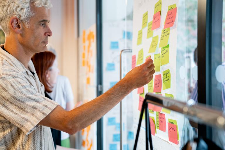 A man with grey hair places a sticky note on a glass wall filled with colourful notes during a brainstorming session.