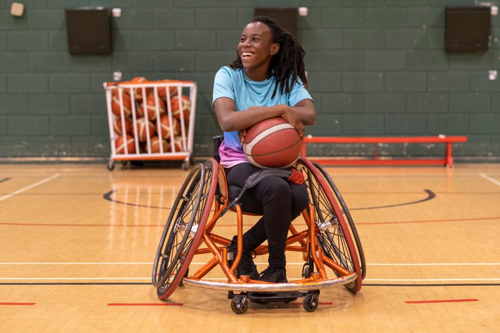 A black young woman in a wheelchair is laughing and holding a basketball