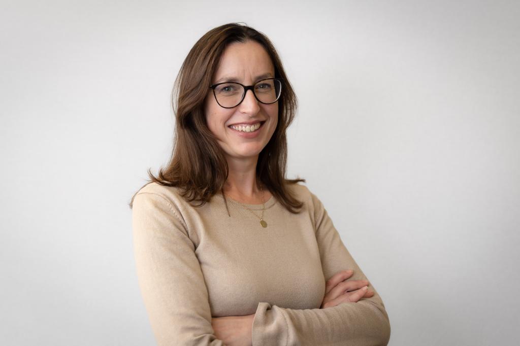 Vicki smiling at the camera. She's a white woman with shoulder-length brown hair, wearing a beige jumper and black rimmed glasses