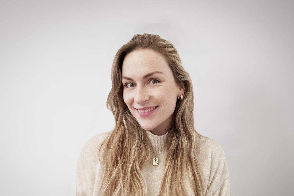 Alexandra smiling at the camera. She's a white woman with long blonde hair, wearing a beige turtle neck jumper and gold earrings.
