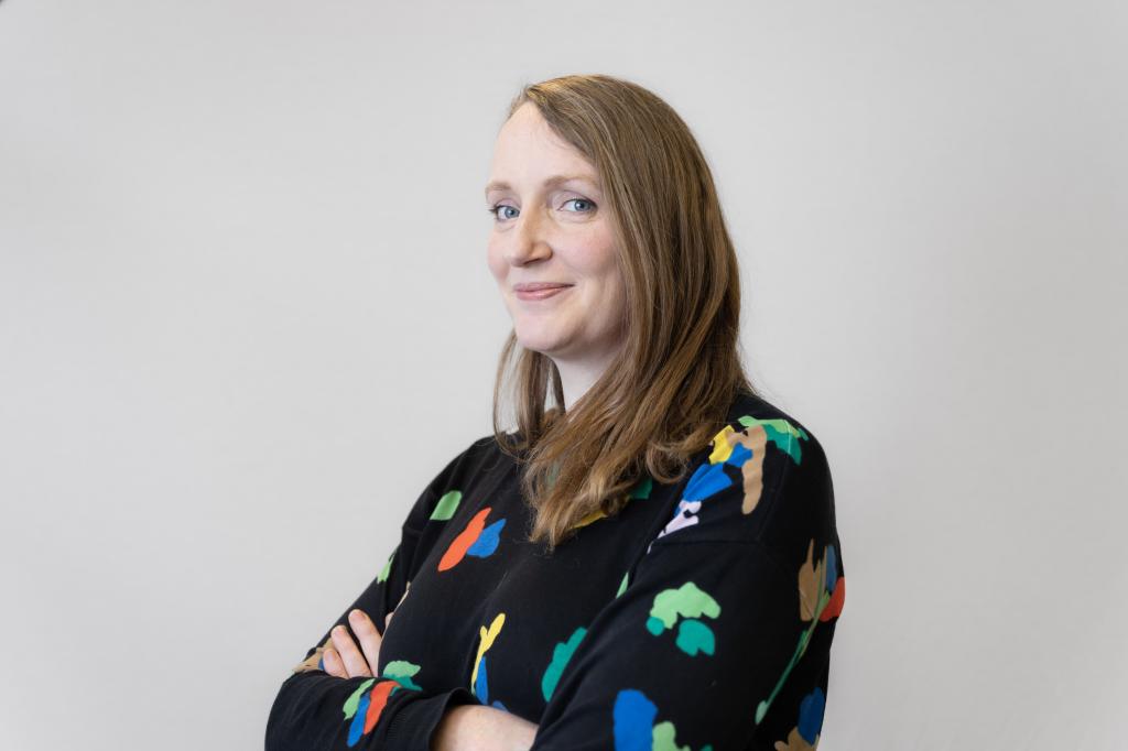 Anna smiling at the camera. She's a white woman with blue eyes and dark blonde hair, wearing a black jumper with colourful shapes on it
