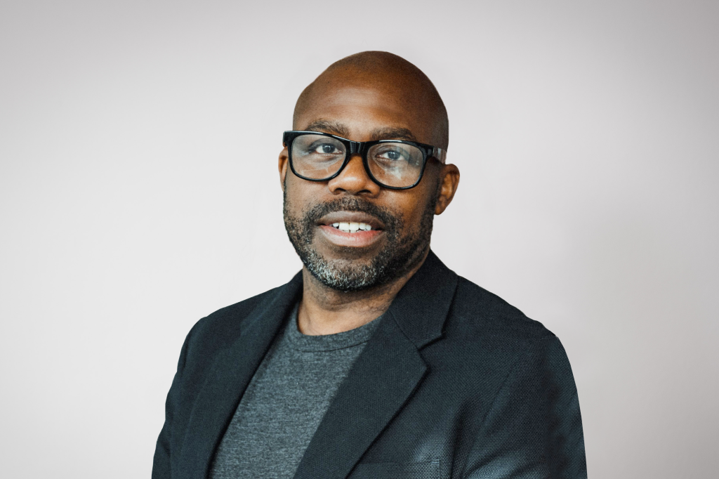 Shane smiling at the camera. He's a bald black man with a short beard, wearing black rimmed glasses with a grey t-shirt and a black jacket
