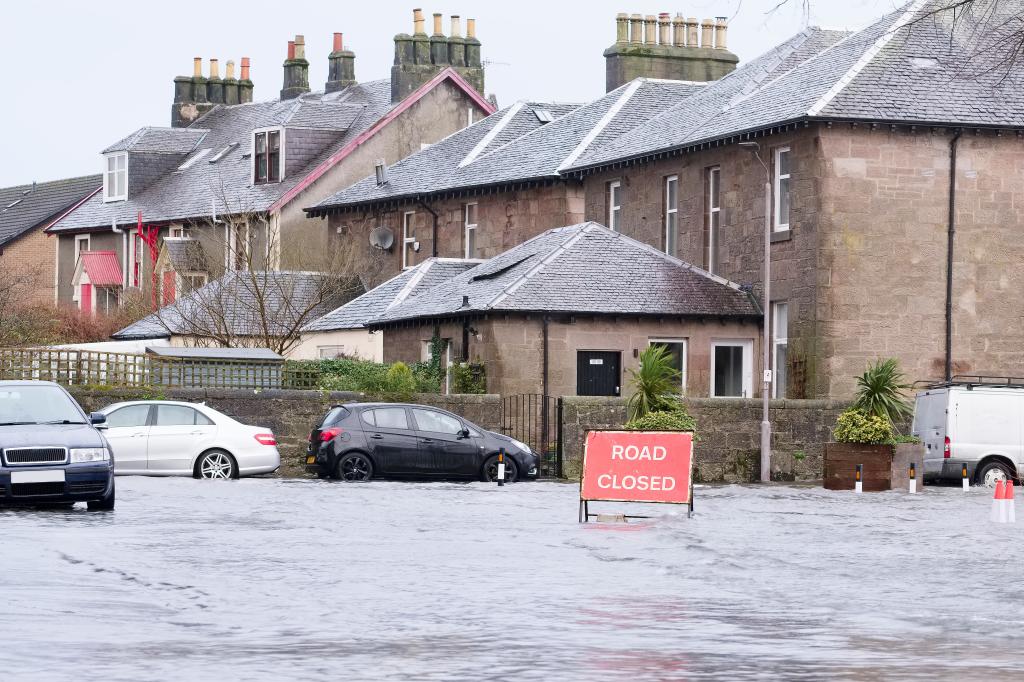 A street that has been flooded, in the background we can see houses and cars that are in floodwater and in the water there is a 'road closed' sign. 