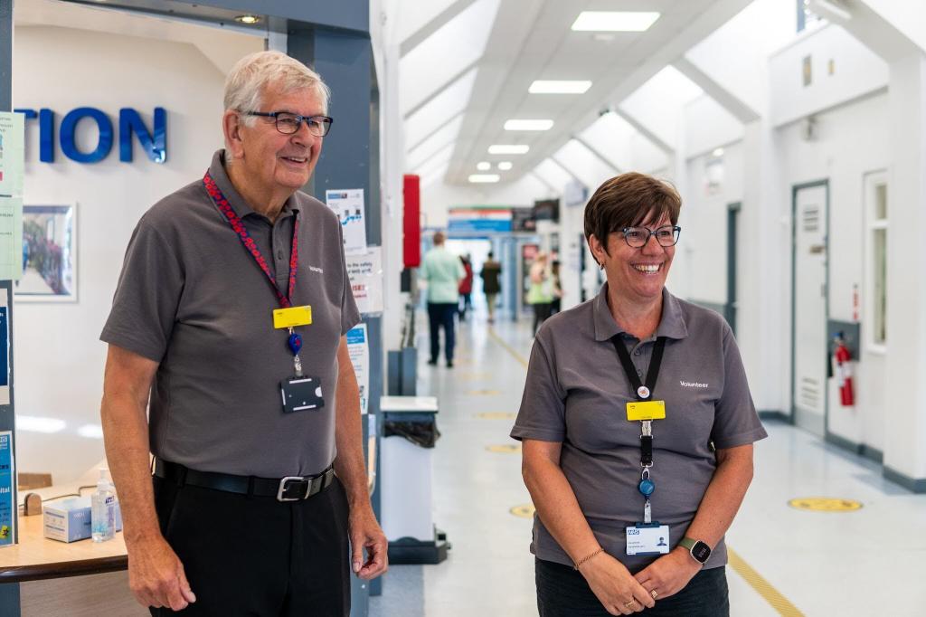 A man and a woman are stood at the reception of a hospital, they are both wearing grey shirts, glasses and lanyards with yellow badges. They are smiling and looking beyond the camera. 