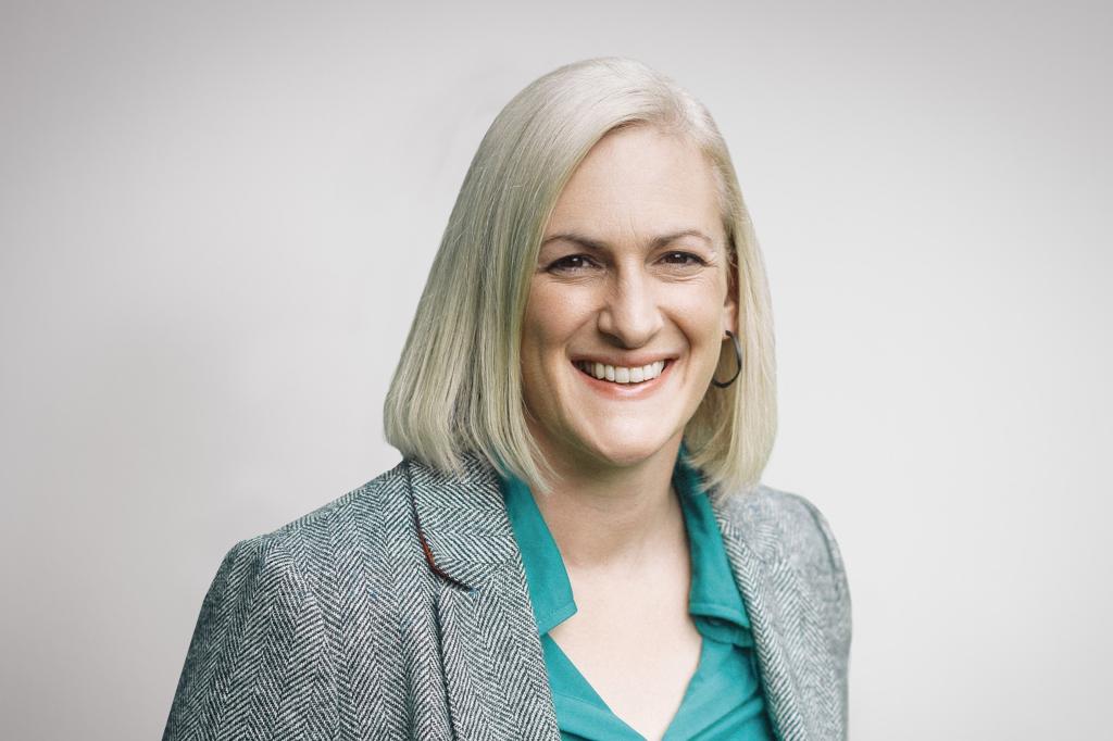 A woman wearing a blue blouse and black and white blazer is smiling into the camera, she has a short haircut. Behind her is a white background.  