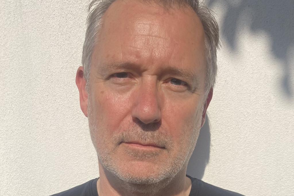 A man wearing a black t-shirt is looking directly at the camera, he has a grey beard and grey hair