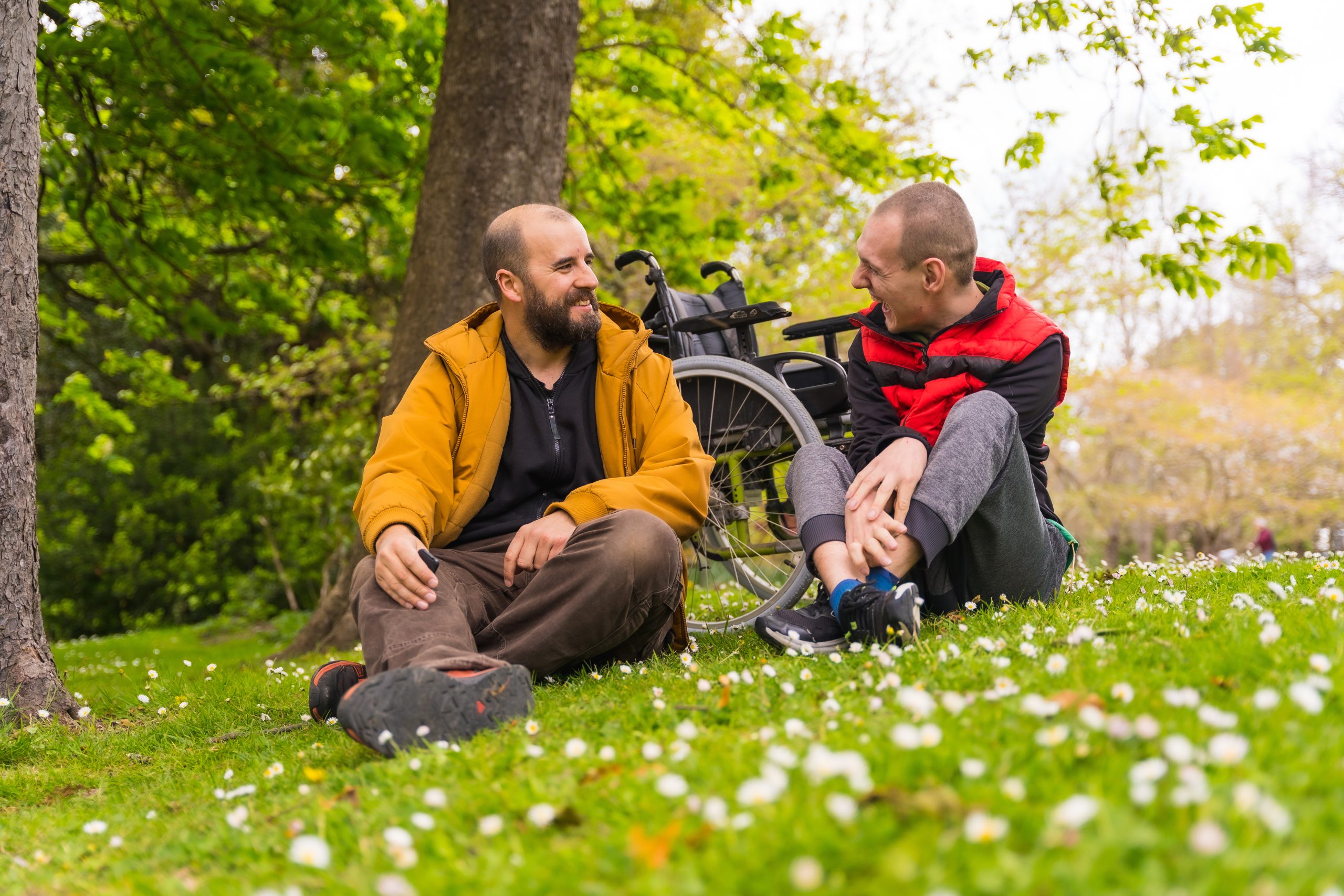 A person with a disability with a friend sitting on the grass of a public park in the city, talking and laughing